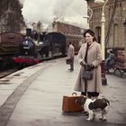 Eine Frau mit Hund steht auf einem Bahnsteig, im Hintergrund fährt ein alter Zug. Menschen sitzen auf einer Bank.