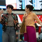 Zwei Personen vor einer Anzeigetafel mit Koffern, „GATWICK EXPRESS“ im Hintergrund. Links kurze Haare, rechts hochgesteckt.