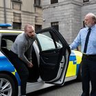 Der Text "Garda" erscheint auf einem Polizeiauto. Ein Mann in Handschellen steigt aus dem Auto, ein Polizist steht daneben.