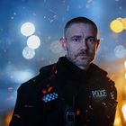 Der Text "POLICE" ist auf einer Uniform sichtbar. Ein Mann steht in einer dunklen Umgebung mit Funken und Lichtern im Hintergrund.