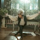 Ein junger Mann in historischer Kleidung posiert dramatisch; im Hintergrund spielen Musiker. Blumenarrangement und Kerzenhalter.