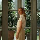 Eine Frau in heller Kleidung steht vor großen Fenstern mit blühenden Pflanzen im Hintergrund.