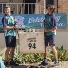 "Ashford Class of 94". Zwei junge Männer in Schuluniformen halten ein Schild, im Hintergrund ist ein Banner sichtbar.