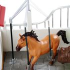 Das Bild zeigt ein Spielzeugpferd in einem kleinen Stallbereich mit einer Sattelbank.