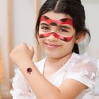 Ein Mädchen mit Marienkäfer-Gesichtsfarbe und -Tattoo lächelt, trägt ein weißes Kleid mit Blütenmuster.