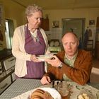 Ein älterer Herr sitzt am Küchentisch, mit einem Teller in der Hand. Eine ältere Dame steht daneben und reicht ihm Essen. Die Küche ist rustikal eingerichtet. Auf dem Tisch liegt Brot.