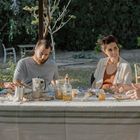 Vier Personen sitzen an einem gedeckten Tisch im Garten. Sonnenlicht fällt auf sie, Bäume im Hintergrund.