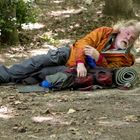 Ein Mann mit orangefarbener Jacke liegt erschöpft auf Waldboden neben einer zusammengerollten Isomatte und einem Rucksack.