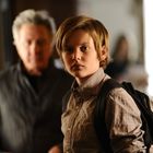 Ein Junge mit Rucksack schaut ernst. Ein älterer Mann unscharf im Hintergrund. Innenraum mit warmer Beleuchtung.