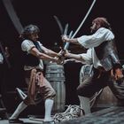 Zwei Männer mit Säbeln duellieren sich auf einem Schiff, umgeben von Fässern und Seilen. Eine Frau beobachtet sie von einer Treppe.