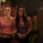 Zwei lächelnde, nasse Frauen stehen draußen im Regen. Eine trägt ein T-Shirt mit lesbarem Text. Im Hintergrund bunte Pflanzen.