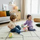 Zwei Kleinkinder sitzen auf einem Teppich und spielen mit bunten Bällen in einem wohnlich eingerichteten Raum.