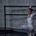 Eine Ballerina in weißem Trikot und rosa Tutu dehnt sich an einer Ballettstange vor einer grauen Ziegelwand.