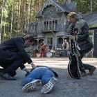 Ein Mann kniet neben einer Person auf dem Boden, ein anderer hält eine Gitarre. Im Hintergrund ein altes Haus im Wald.