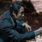 Ein Mann in regennassem Anzug kniet und zeigt mit Nachdruck nach rechts, im Hintergrund verschwommenes Licht.