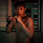 Ich kann den Text nicht erkennen. Ein junger, verschwitzter Mann steht kämpferisch da, mit freiem Oberkörper, vor einem Fenster.