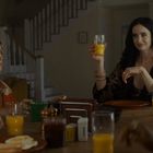 Am Tisch sitzen Menschen beim Frühstück; eine Person hebt ein Glas Orangensaft, andere essen Spiegeleier und Toast.