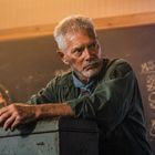 Eine Tafel zeigt ein Punktesystem, daneben ein älterer Mann mit grauem Haar in einem Raum mit Dartscheibe.