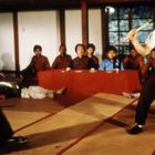 Zwei Männer kämpfen in traditioneller Kleidung, umgeben von sitzenden Zuschauern in einem Raum mit Tatami-Matten.
