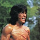 Ein muskulöser Mann mit langen Haaren und blutigen Kratzern auf der Brust, schreit vor einem verschwommenen Hintergrund aus Bäumen.