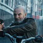 Ein Mann auf einem Motorrad zielt mit einer Pistole im Regen. Im Hintergrund eine Brücke und ein Kanal in einer Stadt.