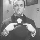 Ein Mann in formeller Kleidung hält einen Melonenhut, trägt weiße Handschuhe und steht vor einem schlichten Hintergrund.