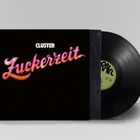 "Cluster" in Weiß und "Zuckerzeit" in rosa, geschwungener Schrift mit Glanzeffekten auf schwarzem Hintergrund. Schallplatte.