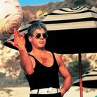Ein Mann in einem schwarzen Tanktop dreht ein großes, spiralförmiges Objekt auf seinem Finger. Im Hintergrund Strandschirme.