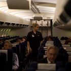 Eine Flugbegleiterin steht im Gang eines Flugzeugs, umgeben von sitzenden Passagieren. Es herrscht gedämpftes Licht.
