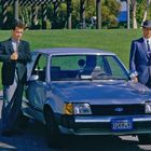 Kennzeichen: "CALIFORNIA 1PCE253". Zwei Männer in Anzügen stehen neben einem blauen Auto im Freien.