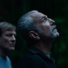 Zwei Männer blicken nach oben, einer im Vordergrund mit grauem Haar, der andere im Hintergrund, dunkel bewaldeter Hintergrund.