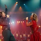 Zwei Personen singen mit Mikrofonen auf einer Bühne, umgeben von bunten Lichtern und einem Blasorchester im Hintergrund.