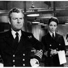 Person in Marineuniform, ernst blickend, neben einer anderen Person, die Notizen macht, in einem Büro mit alter Ausstattung.