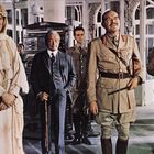 Fünf Männer in historischer Kleidung, darunter ein Beduinengewand und britische Militäruniformen, stehen in einem eleganten Raum.