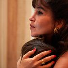 Eine Frau im Profil mit langen braunen Haaren und roten Fingernägeln umarmt ein Kind. Sie trägt goldene Ohrringe.