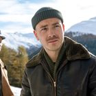 Zwei Männer im Winter: Der vordere trägt eine grüne Mütze und Lederjacke, der hintere einen Hut und Mantel, vor Bergkulisse.