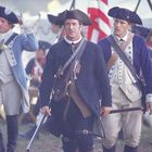 Drei Männer in historischen Militäruniformen, zentrale Figur trägt ein Gewehr, ernstes Gesicht, Hintergrund mit Zelten.