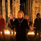 Fünf Personen in dunkler Umgebung, umgeben von hohen, dekorativen Holzkonstruktionen und einem Kreis aus Kerzen.