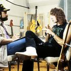 Zwei Männer sitzen in einem Raum voller Gitarren und unterhalten sich. Einer trägt eine Mütze mit Text.