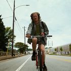 Eine Person mit Kopfhörern fährt auf einer leeren Straße Fahrrad. Stromleitungen verlaufen parallel dazu.