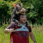 Ein Mann mit einem Wildschwein auf den Schultern steht in einem grünen, grasbewachsenen Gebiet.