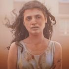 Eine Frau mit welligem Haar steht vor unscharfem Hintergrund, wirkt erschöpft, ihr Gesicht ist leicht schmutzig.