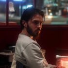 Ein Mann mit Bart sitzt im Innenraum, umgeben von dunklen Tönen und einem leuchtenden Heizgerät im Hintergrund.