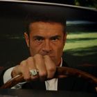 Ein Mann mit ernstem Ausdruck sitzt am Steuer eines Autos, trägt Anzug und auffälligen Ring.