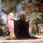 Ein dunkles Wesen in majestätischem Gewand steht vor einem großen, rosa Kristall in einer mystischen, höhlenartigen Umgebung.