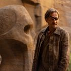 Ein Mann mit Augenklappe in felsiger Landschaft, steht neben großer Schädelstatue. Er trägt braune Jacke.