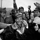 Eine Gruppe fröhlicher Menschen umarmt einen Mann in Fliegerkleidung mit einer Zigarette und Fernglas. Alle lächeln.