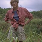 Ich kann keine bestimmten Texte erkennen. Eine blonde Frau steht in hohem Gras, trägt ein rotes Hemd und beige Shorts.