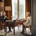 Zwei Frauen sitzen sich an einem Holztisch gegenüber, der Raum hat große Fenster mit Vorhängen und ist gemütlich eingerichtet.