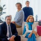 Vier Personen sitzen auf einem Sofa. Einer trägt Streifen, eine Frau trägt Schals in Blau. Pflanzen und Deko im Hintergrund.